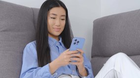 Asian girl using mobile phone. Cheerful Vietnamese female browsing smartphone app while sitting on couch at home. BIPOC person lifestyle video clip