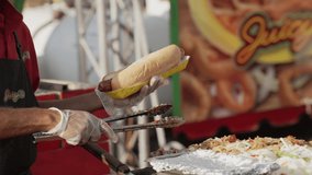 This video shows a close up view of a food vendor adding fresh onions and peppers with tongs to large hotdogs.