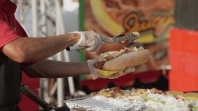 This video shows a close up view of a food vendor adding fresh onions and peppers with tongs to large hotdogs.
