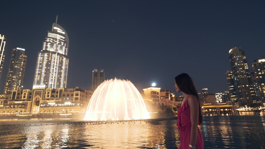 Dubai, UAE-10.10.2021:Tourist young woman walking along the Dubai Fountain during Dubai Dancing Fountain Water Show at night. Woman enjoying the view of Dubai Fountain Water Show