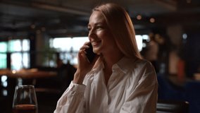 Smiling pretty blonde young woman talking on mobile phone sitting at table with red wine in modern restaurant with dark interior. Attractive happy female having conversation on smartphone in bar. - Powered by Shutterstock - Get 15% off with code: PIKWIZARD15