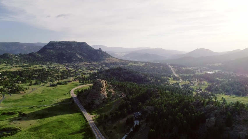 Drone pan from right to left in the golden hour of the morning in Estes Park Colorado. Mountains casting shadows on the field, sun on the right, peaks in the distance and a lake in left foreground.