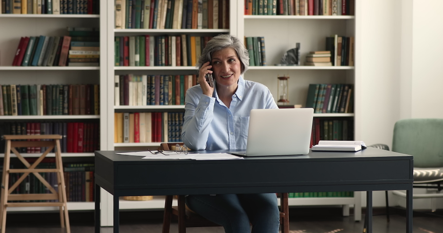 Happy positive senior 60s business woman talking on mobile phone at workplace table, sitting at laptop, speaking, listening, smiling, laughing, discussing work, making call. Communication concept Royalty-Free Stock Footage #1093573477