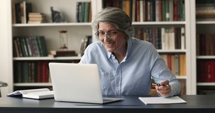 Senior grey haired college teacher talking to student, making video call, speaking at webcam, using laptop, giving feedback to essay, reviewing paper documents on table
