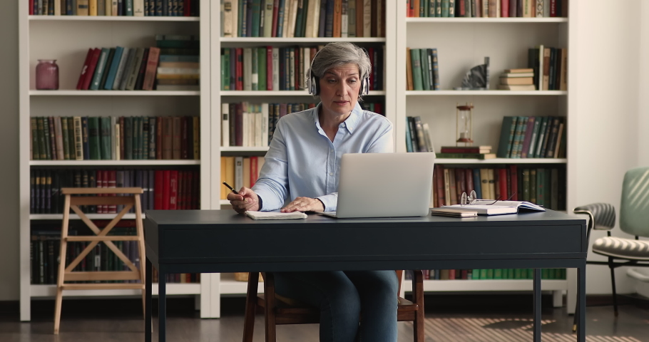 Focused senior student woman wearing wireless headphones talking to teacher on video call, using laptop for chat, virtual conference, online learning, studying, writing notes Royalty-Free Stock Footage #1093573533