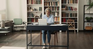 Serious older mature business woman working in office, using laptop, typing at table with papers, bookshelves behind Reading text, watching video, thinking over project
