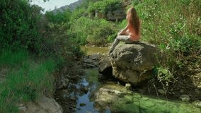 Woman is sitting on a stone and enjoying the nature. Beautiful landscape with a small lake. Green pond and stones. Sunny nice weather. Blue sky. Horizontal video. 