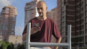 Moving camera. bald man with a cigarette trains on a stationary bike in a city park in casual clothes.Talking on the phone on video call.Street workout.Wrong lifestyle,the harm of smoking