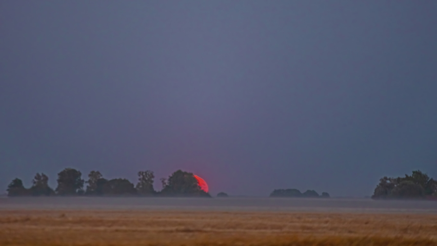 Super moon rises over a farm field with a low layer of fog or mist to glow over the countryside - time lapse