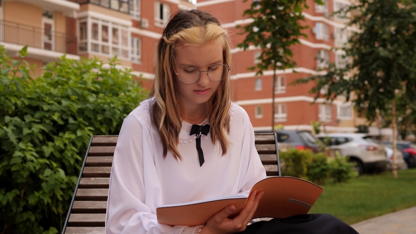 A smart teenage girl in a white school blouse and glasses reads notes in a notebook in the yard sitting on a bench after school.