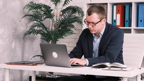 Slow motion of the side view of a serious businessman in glasses and a suit sitting in the office at the desk and printing something on a laptop