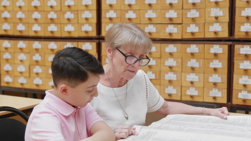 4K. A happy elderly grandmother listens to a little school-age boy reading a book in the library.