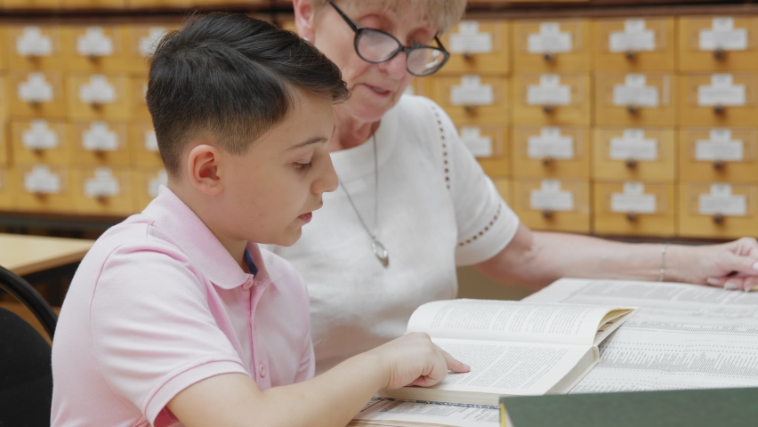 4K. A happy elderly grandmother listens to a little school-age boy reading a book in the library.