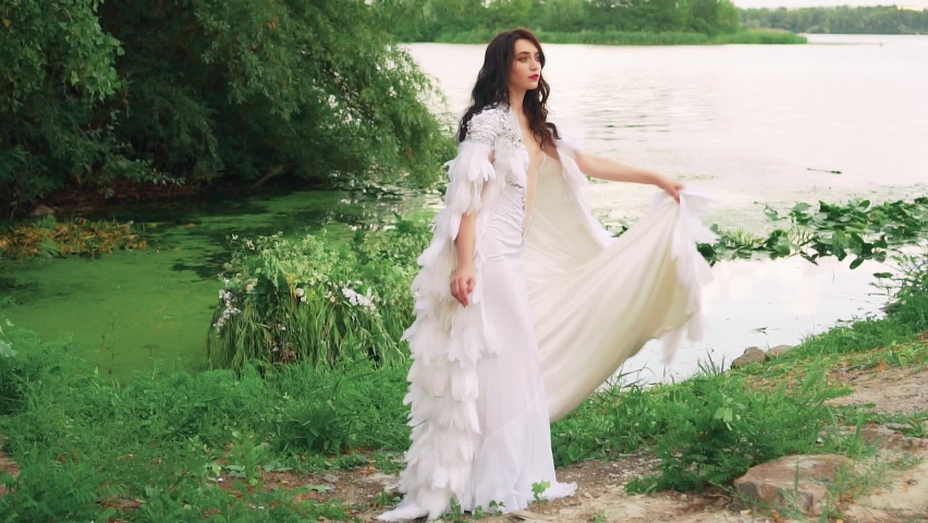 Fantasy woman sexy queen in long white dress stands fashion model posing by lake. Creative clothing, cloak cape with natural feathers. Summer nature water river green trees. Girl princess beauty