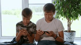 Two boys play computer games at home on their smartphones, watch videos, rejoice and laugh. Two brothers watch interesting videos together on tablet or smartphone. They laugh and indulge. Sit on table