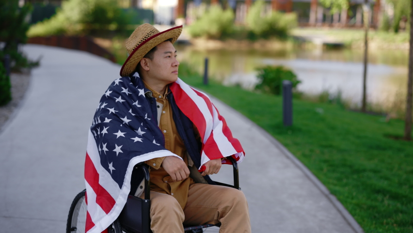 Portrait of the veteran sitting on the wheelchair carying the usa flag in the middle of the street near the local lake. Close view of the serious soldier resting in summer evening in the street
