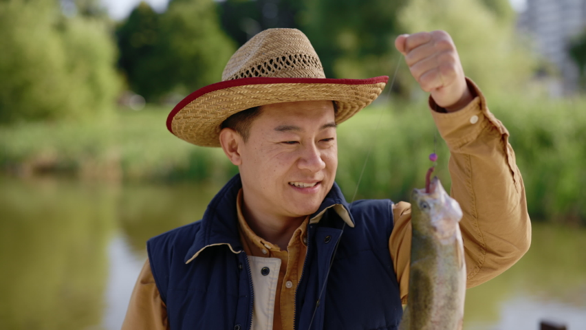 Close view of the asian man wearing special fishing clothes holding the fish he caught on the hook and feeling happy about it looking at the camera