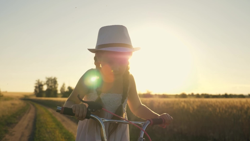 A girl in a cowboy hat rides a bicycle on the road in a field. The kid is enjoying the meadow landscape at sunset. The baby rides a bike and laughs. The child is dreaming.