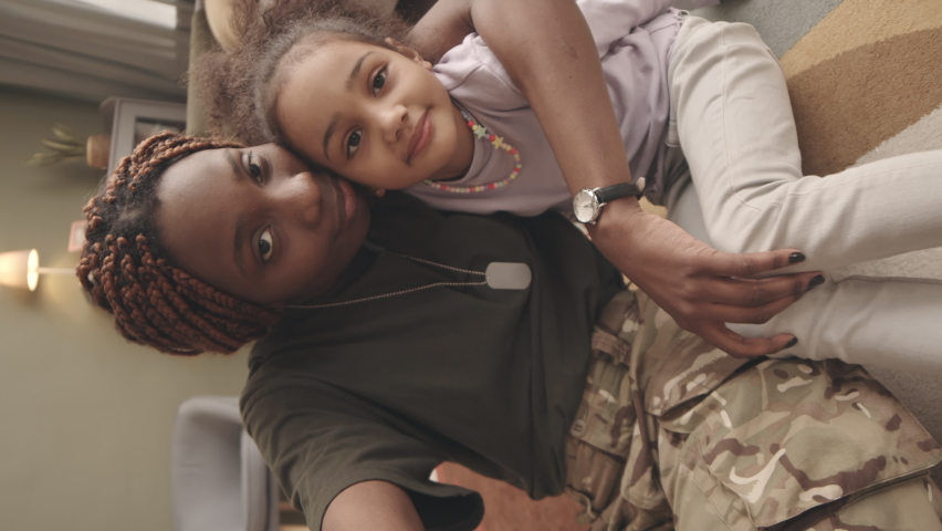 Vertical handheld POV slowmo of young African American military woman and her cute little daughter showing v signs and smiling at camera while taking selfie portrait, sitting on rug in living room