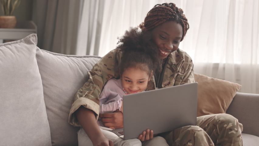 Medium slowmo of loving African American mother in military uniform and her little daughter smiling and embracing on sofa, watching funny videos on laptop during leisure time Royalty-Free Stock Footage #1093626769
