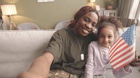 Handheld POV slowmo of young Black woman soldier and her little daughter with US flag in hands smiling at camera while taking selfie portrait, sitting on soft grey couch in living room - Powered by Shutterstock - Get 15% off with code: PIKWIZARD15