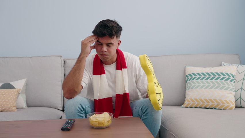 Young hispanic man supporting soccer team sitting on sofa at home