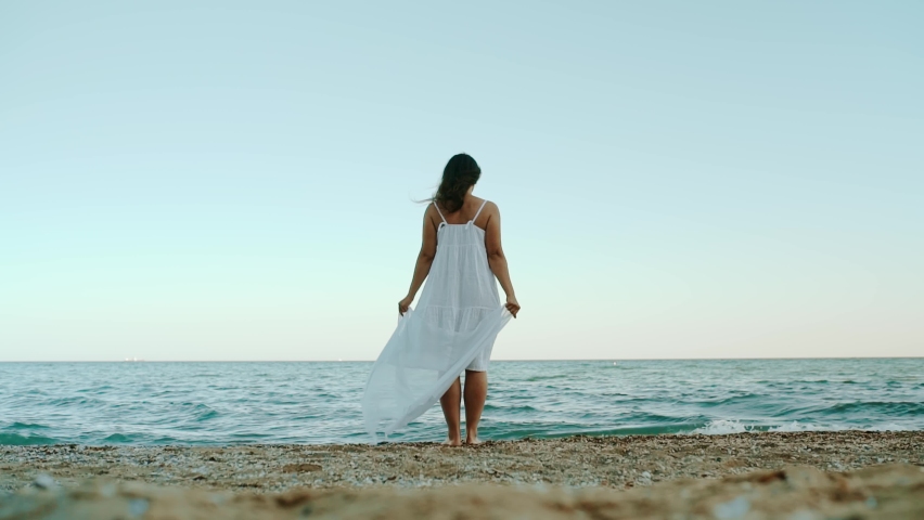 Young woman in a white dress stands on the seashore and looks into the distance