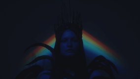 Zoom out portrait shot of woman wearing gothic crown and feather dress posing for camera in dark studio with blue neon light and rainbow projection - Powered by Shutterstock - Get 15% off with code: PIKWIZARD15