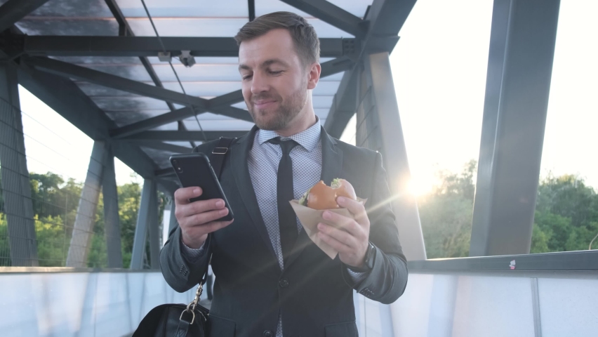 A businessman walking down the street and eating fast food. 