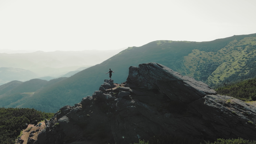 A man standing on top of a mountain with and raising his hands up the mountain. Victory and overcoming the top. A tourist looking at the sunrise on top of a rock.