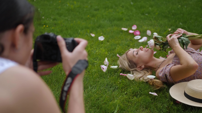 photographer takes pictures of a girl lying a beautiful woman in a green meadow, who holds a bouquet of peonies in front of her face in summer in nature