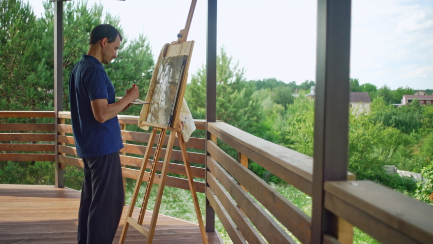 A male artist stands on the terrace at home and paints a picture with a brush. The art and hobby of easel painting. High quality 4k footage