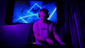 DJ in club working indoors in neon light. Caucasian male deejay person in nightclub using headphones, laptop and control console to mix audio music tracks list. Nightlife concept. Slow motion video