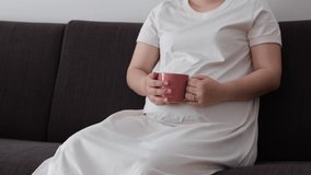 A video of a pregnant woman sitting on a sofa holding a cup and stroking her belly