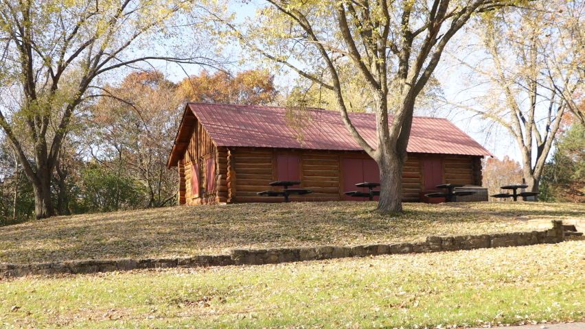Log built park shelter on a beautiful sunny autumn day.  Leaves all over the grass. Shadows cast from the tree branches. Blue sky and green grass. Red metal roof on building. 