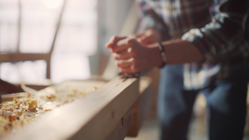 Close Up Portrait of a Handsome Young Artisan Craftsman in Checkered Shirt Using Hand Plane to Shape a Wood Bar. Carpenter Working on a Project in a Loft Studio with Tools on Walls.