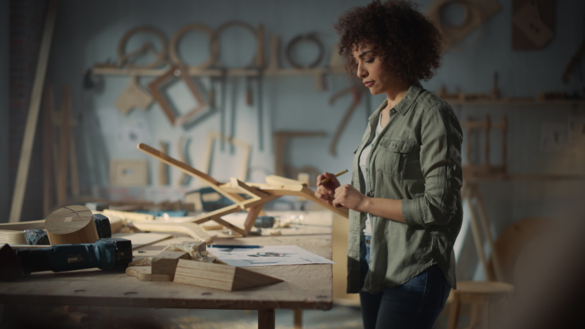 Portrait of a Young Beautiful Carpenter Checking a Blueprint and Starting to Assemble a Wooden Chair. Professional Furniture Designer Working in a Studio in Loft Space with Tools on the Walls.
