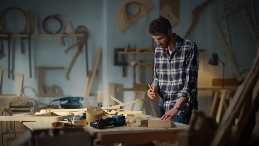 Young Artisan Furniture Designer Marking Out Dimensions on a Blueprint and Starting to Assemble a Wooden Chair. Hipster Carpenter Working in a Studio in Loft Space with Tools on the Walls.
