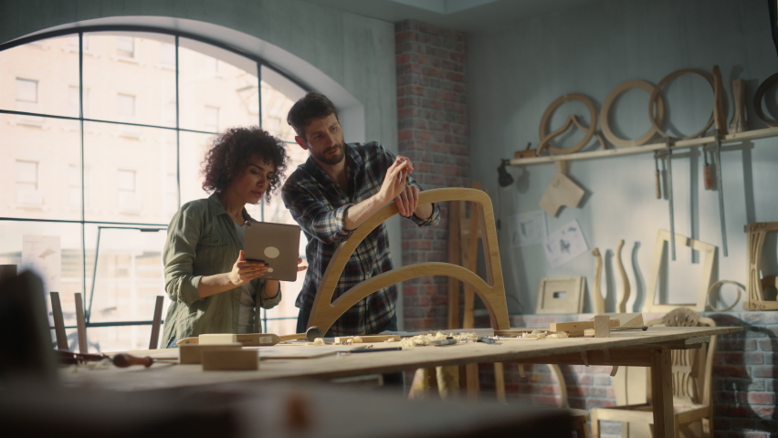 Two Talented Small Business Owners Using Tablet Computer and Discussing the Design of a New Wooden Chair in a Furniture Workshop. Carpenter and a Young Female Apprentice Working in Loft Studio. Royalty-Free Stock Footage #1093664179