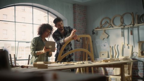 Стоковое видео: Two Talented Small Business Owners Using Tablet Computer and Discussing the Design of a New Wooden Chair in a Furniture Workshop. Carpenter and a Young Female Apprentice Working in Loft Studio.