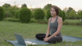 Consultation, yoga video lesson, sports woman instructor actively gestures, teaches yoga to students in front of a laptop while sitting in the park in the evening.