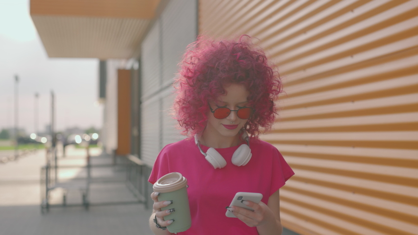 A pink curly teenager is walking and using a smartphone outdoors. Hipster girl is browsing Internet on a phone or texting and chatting. Coffee break. Network and sms or communication concept.