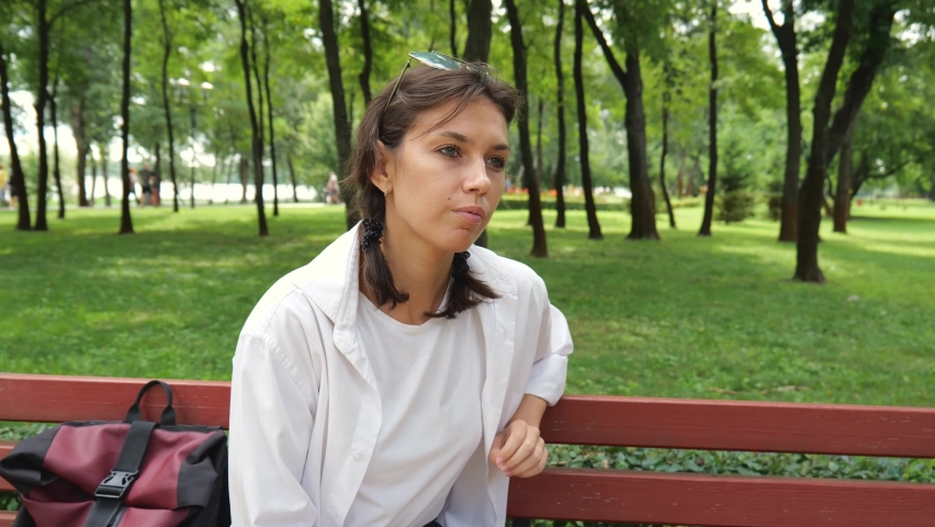 A young brunette woman in a white shirt is sitting on a bench talking on a mobile phone in a city park. Cheerful female tourist