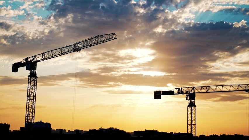 Dark silhouettes working and moving of construction cranes and unfinished residential building on yellow beautiful sunrise and blue sky background. Housing construction, apartment block in city.