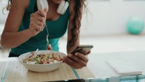 Video of beautiful sporty woman eating a bowl of salad while messaging with her mobile phone in the kitchen at home.