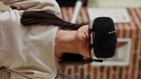 Vertical video: Asian person wearing futuristic VR goggles while interacting with cyber reality. Playful woman wearing virtual reality headset scrolling through metaverse while working from home