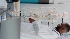 Vertical video: African american osteopath pointing at human skeleton to explain bones disease and diagnosis to senior patient at checkup visit. Anatomy specialist showing spinal cord at osteopathy