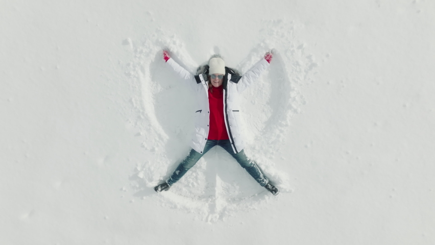 TOP down aerial view 4K footage of Happy woman making snow angel on sunny winter day. Funny charming female laying down on fresh perfect white snow surface looking up in the air. Winter background