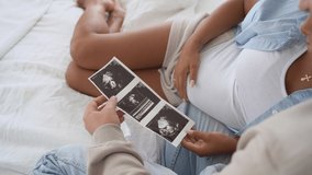 Multiracial pregnant couple watching ultrasound image picture at home. Closeup feeling happy, stroking tummy and excited on seeing baby ultrasound - Powered by Shutterstock - Get 15% off with code: PIKWIZARD15