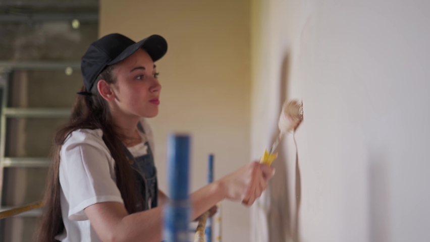 Female painter paints wall with roller on scaffolding at construction site. Young woman in overalls doing home decoration, renovation on staging. Professional worker makes DIY repair work in room. Royalty-Free Stock Footage #1093690271